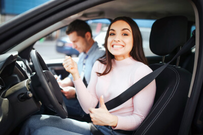 Male auto instructor takes exam in young woman. Happy positive model look on camera with big thumbs up. Smiling with likes. Instructor sit beside her. Passing exams. Daylight outside